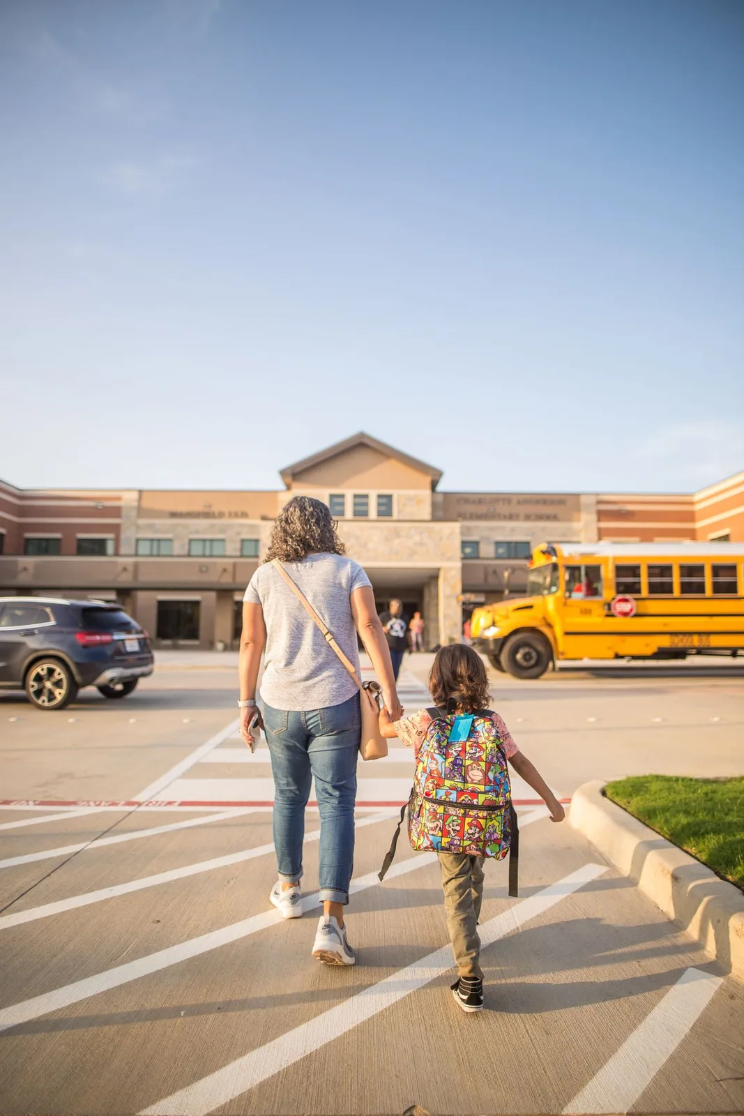 Back view of unrecognizable woman in casual wear holding schoolgirl with backpack by hand while walking on asphalt pavement with marking lines near exterior of building and bus under sky
