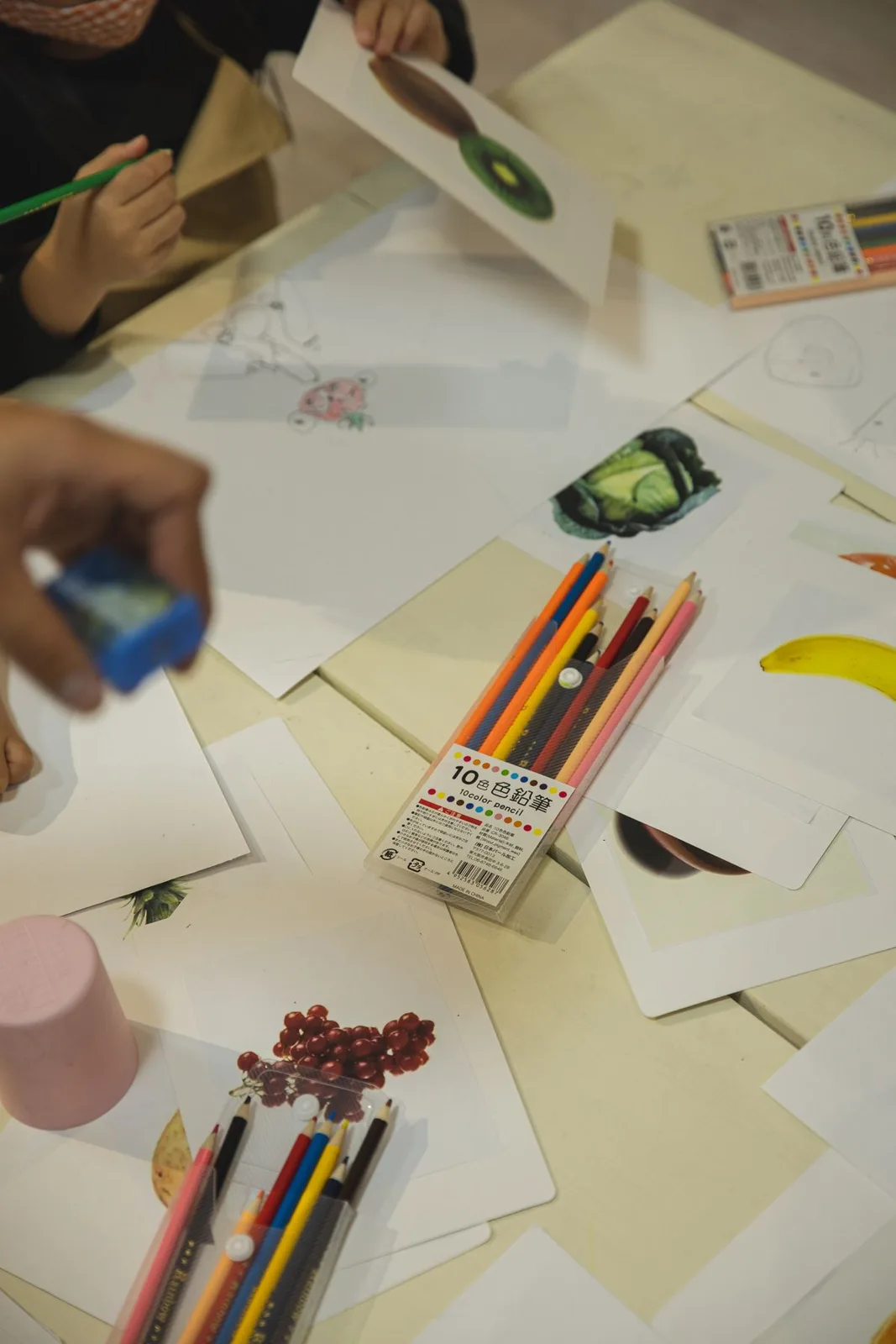 Overhead view of crop anonymous kids sitting at table with drawings on paper sheets and colored pencils