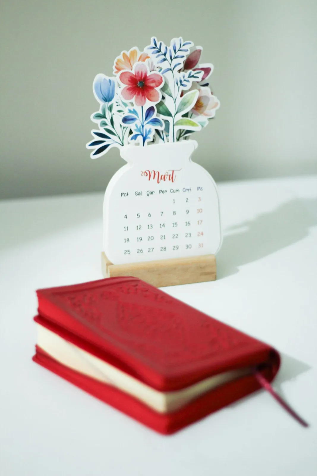 A beautifully designed March calendar with flowers on a white table beside a red notebook.