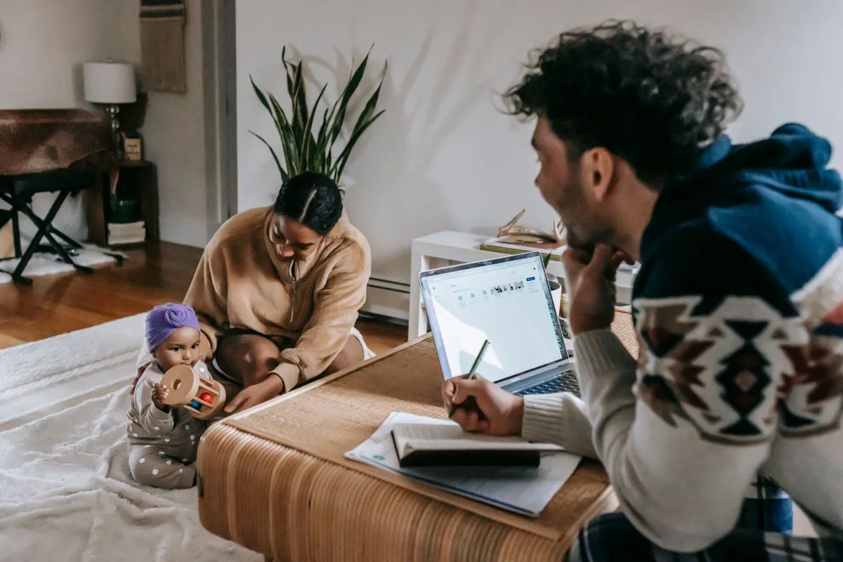Ethnic male taking notes in notepad at table with laptop while working online in living room with African American mother and little toddler
