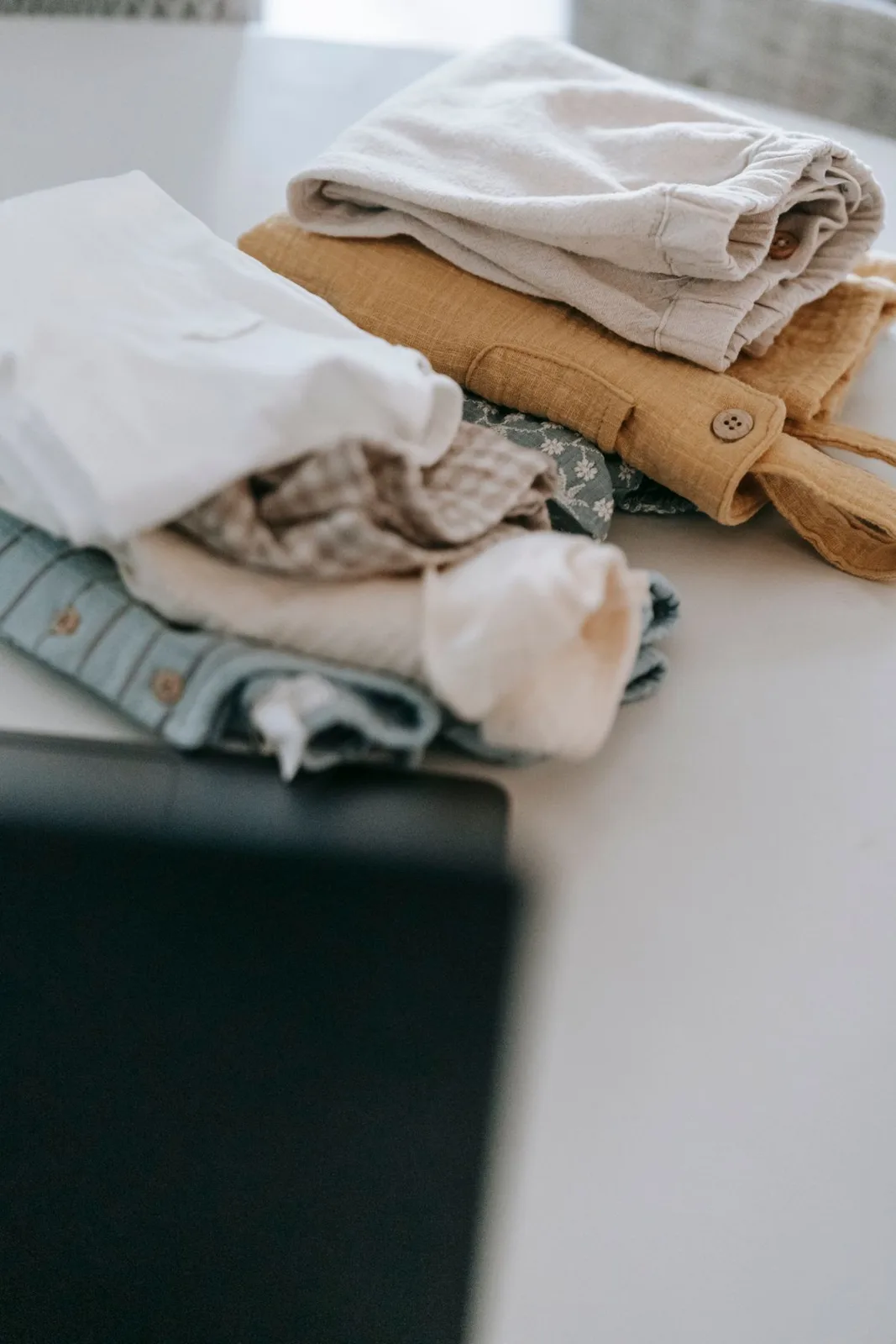 Abundance of colorful baby clothes folded on white clean table with opened modern blurred laptop in light room at home