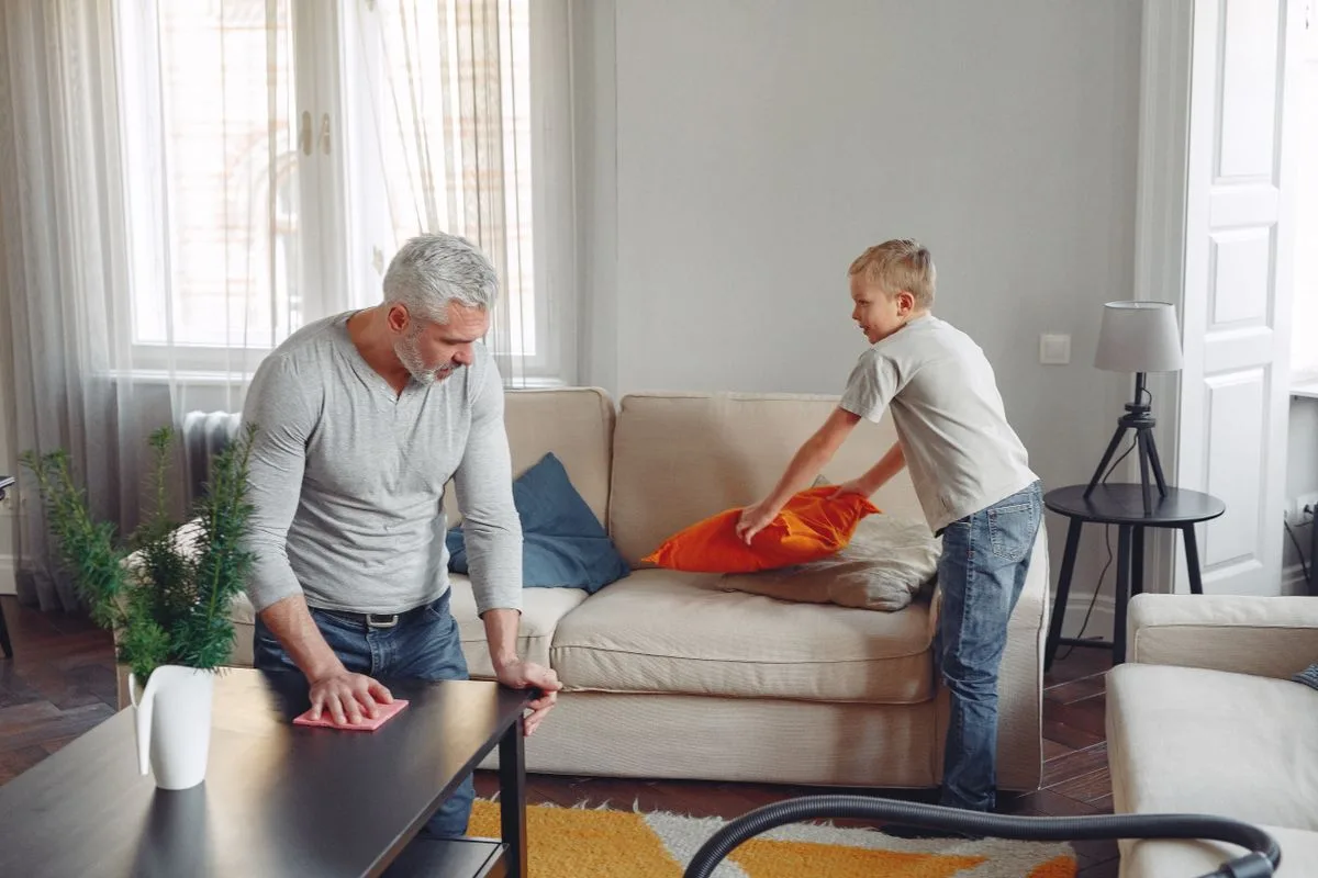 A father and son clean their living room, tidying up cushions and dusting tables.
